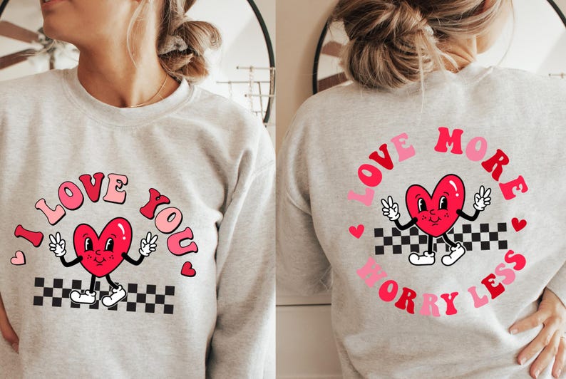 May include: A light gray sweatshirt with a retro design. The front features the text "I LOVE YOU" with a cartoon heart character. The back has "LOVE MORE WORRY LESS" with a similar heart character. The design includes pink, red, black, and white.