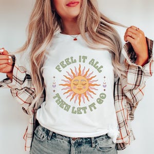 May include: White t-shirt with a graphic of a sun with a face and the text "Feel it all then let it go" in green.