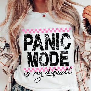 Könnte beinhalten: Weißes T-Shirt mit dem Text "PANIC MODE is my default" in verblichener schwarzer Schrift, umrahmt von einem rosa-weiß karierten Muster. Ein lässiges Kleidungsstück für den Alltag.
