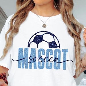 May include: White t-shirt featuring a blue glitter soccer ball design and the words "MASCOT soccer" in blue. The shirt has a casual style and is suitable for everyday wear.