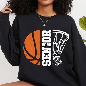 May include: Black sweatshirt with a white basketball and hoop graphic. The basketball is orange and has a distressed look. The text "Senior 2026" is printed vertically on the sweatshirt.