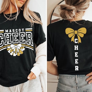 Puede incluir: Camiseta negra con texto en purpurina dorada que dice "MASCOT CHEER" y un lazo de purpurina dorada con pompones blancos. La parte posterior de la camiseta tiene un lazo de purpurina dorada y texto en purpurina blanca que dice "CHEER" verticalmente.