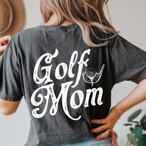 Pu&ograve; includere: Una maglietta nera con una grafica bianca invecchiata che dice "Golf Mom" con una mazza da golf e una palla.