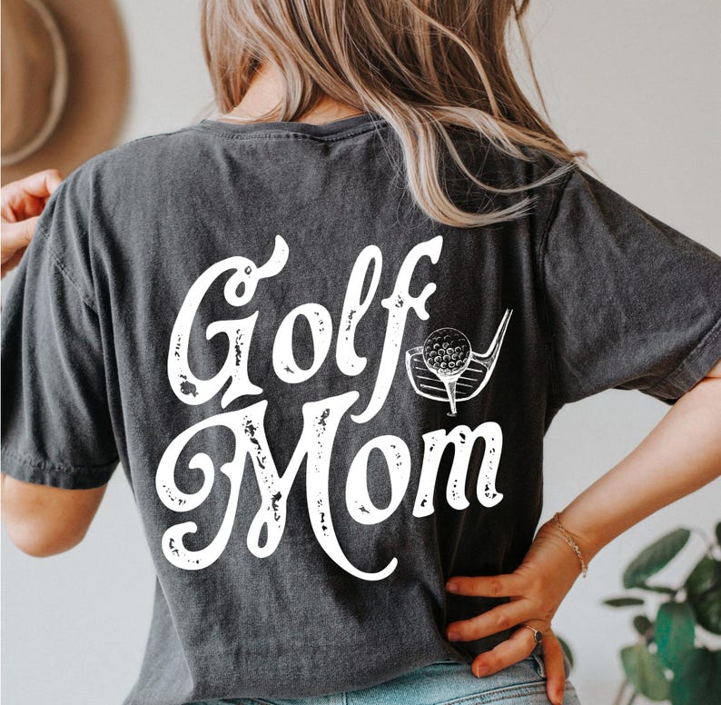 Pu&ograve; includere: Una maglietta nera con una grafica bianca invecchiata che dice "Golf Mom" con una mazza da golf e una palla.