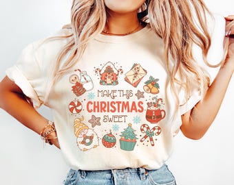 Könnte beinhalten: Cremefarbenes T-Shirt mit einem festlichen Weihnachtsgrafik. Das Design zeigt eine kreisförmige Anordnung von Illustrationen zum Thema Feiertage, darunter ein Lebkuchenmann, Zuckerstangen und der Text "MAKE THIS CHRISTMAS SWEET."