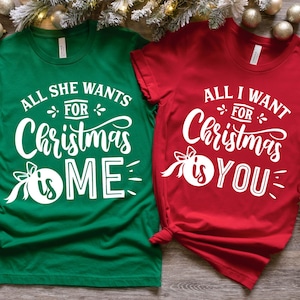 Puede incluir: Dos camisetas de manga corta con temática navideña. Una es verde con el texto "All She Wants For Christmas Is Me" en blanco. La otra es roja con el texto "All I Want For Christmas Is You" en blanco.