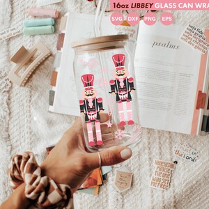 May include: A clear 16oz Libbey glass can wrap with a wooden lid, featuring a design of two toy nutcracker soldiers in red, black, and pink. The words "16oz LIBBEY GLASS CAN WRAP" are at the top. The glass is held in a hand.