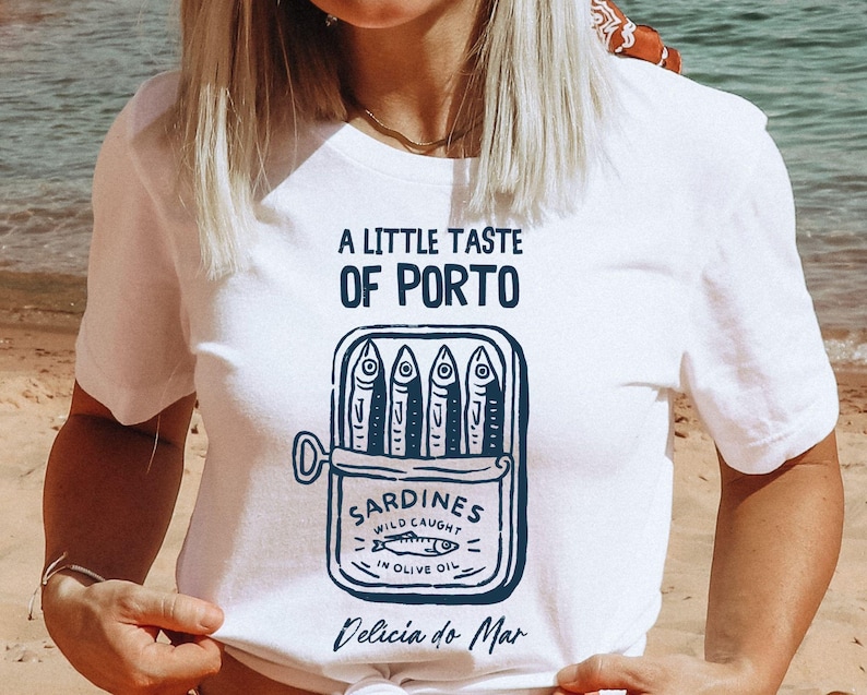 May include: White t-shirt with a navy blue graphic of a sardine can, featuring the text "A Little Taste of Porto" and "Sardines Wild Caught in Olive Oil". The phrase "Delicia do Mar" is also printed on the shirt.