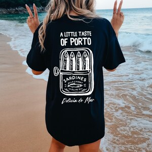 May include: Black t-shirt with a white graphic of a sardine can and the text "A Little Taste of Porto" and "Delicia do Mar". The sardine can reads "Sardines Wild Caught in Olive Oil".