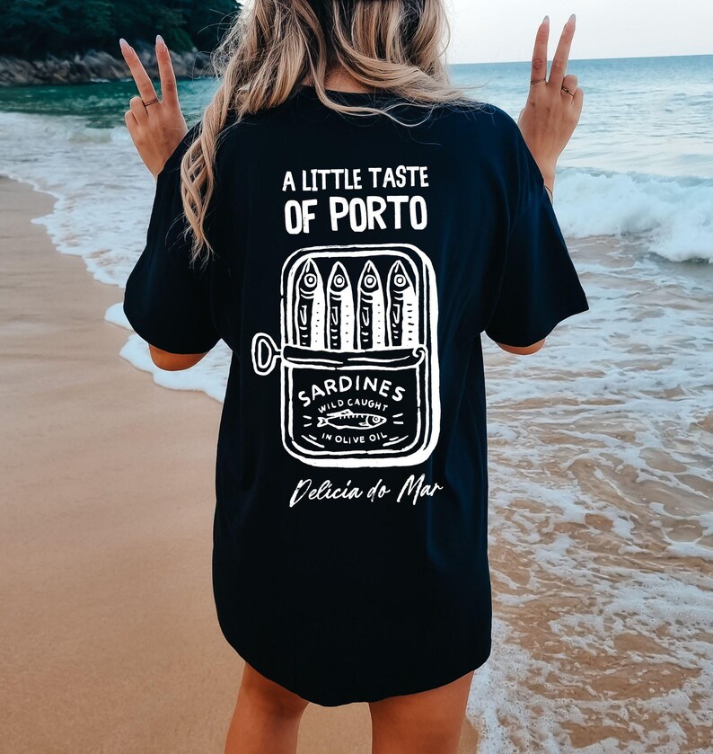 May include: Black t-shirt with a white graphic of a sardine can and the text "A Little Taste of Porto" and "Delicia do Mar". The sardine can reads "Sardines Wild Caught in Olive Oil".