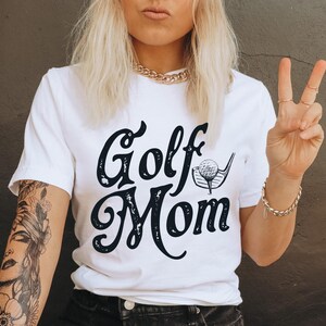 Pu&ograve; includere: Maglietta bianca con testo nero che dice "Golf Mom" e una grafica di una mazza da golf e una palla.