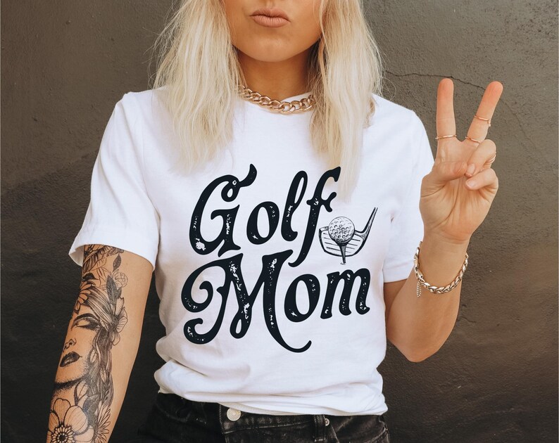Pu&ograve; includere: Maglietta bianca con testo nero che dice "Golf Mom" e una grafica di una mazza da golf e una palla.