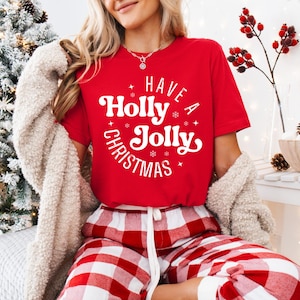 Puede incluir: Camiseta roja con el texto "Have a Holly Jolly Christmas" en blanco, combinada con pantalones de pijama a cuadros rojos y blancos. Un cárdigan color crema está drapeado sobre los hombros. La escena está ambientada con un árbol de Navidad y decoraciones festivas.