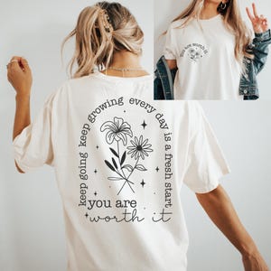 May include: A white t-shirt with a black floral design and the text "keep going keep growing every day is a fresh start you are worth it" on the back. The front of the shirt has the text "you are worth it" with a small floral design.