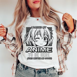 Op de afbeelding: Wit T-shirt met een zwart-witte afbeelding van een anime-personage. De tekst op het shirt luidt "Ik heb mijn anime gepauzeerd om hier te zijn (dit moet wel goed zijn)".