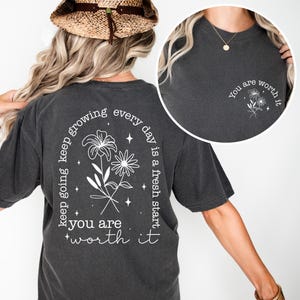 May include: A charcoal gray t-shirt with a white floral design and the text "keep going keep growing every day is a fresh start you are worth it" on the back. The front of the shirt has a smaller white floral design and the text "You are worth it".