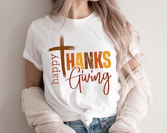 May include: White t-shirt with a Thanksgiving-themed design. The design features a brown cross, the words "happy" in vertical brown text, and the words "THANKS GIVING" in orange and brown text. The shirt is tied at the waist.