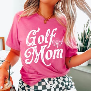 Pu&ograve; includere: Una maglietta rosa con il testo "Golf Mom" in bianco con un aspetto usurato. Il testo &egrave; in un font corsivo con un grafico di una mazza da golf e una palla accanto.