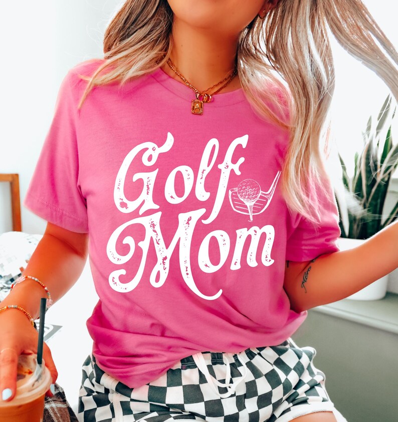 Pu&ograve; includere: Una maglietta rosa con il testo "Golf Mom" in bianco con un aspetto usurato. Il testo &egrave; in un font corsivo con un grafico di una mazza da golf e una palla accanto.