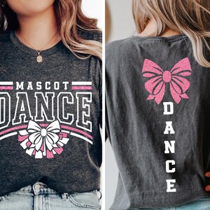 May include: A dark grey t-shirt with a white and pink glitter design. The front of the shirt says "MASCOT DANCE" with a pink bow and white pom-poms. The back of the shirt has a pink bow and the word "DANCE" spelled vertically in white.