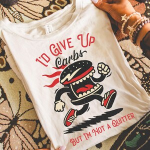 Puede incluir: Camiseta color crema con un personaje de hamburguesa de dibujos animados corriendo. El texto "I'D GIVE UP CARBS" y "BUT I'M NOT A QUITTER" están impresos en rojo. La hamburguesa tiene un pan negro, boca roja y zapatillas rojas.