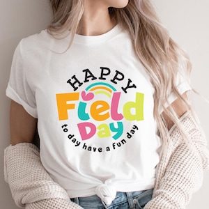 Könnte beinhalten: Weißes T-Shirt mit einem farbenfrohen "Happy Field Day"-Aufdruck. Das Design beinhaltet einen Regenbogen, Herzen und die Sätze "to day" und "have a fun day" in einer verspielten Schriftart. Das Shirt besteht aus einem weichen Material.