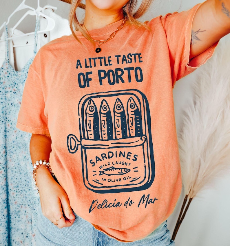 May include: Orange t-shirt with the text "A Little Taste of Porto" and a sardine can graphic. The graphic includes the words "Sardines Wild Caught in Olive Oil" and "Delicia do Mar".