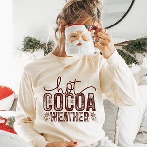 Puede incluir: Camiseta de manga larga color crema con las palabras "hot COCOA WEATHER" en una fuente decorativa de color rojo oscuro. La camiseta presenta detalles de copos de nieve. Una taza de Papá Noel se sostiene frente a la cara.