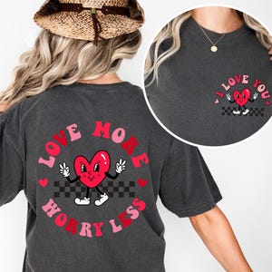 May include: Dark gray t-shirt with a graphic of a red heart character with peace signs and the text "LOVE MORE WORRY LESS" in a circular design. A smaller version of the heart graphic is on the upper chest with the text "I LOVE YOU".