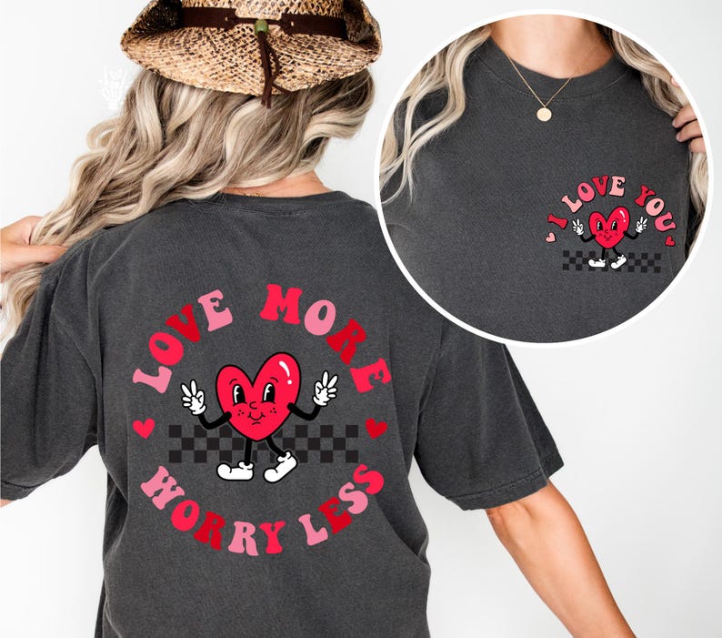 May include: Dark gray t-shirt with a graphic of a red heart character with peace signs and the text "LOVE MORE WORRY LESS" in a circular design. A smaller version of the heart graphic is on the upper chest with the text "I LOVE YOU".