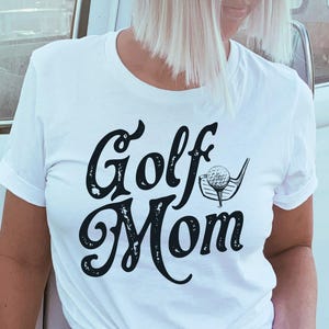 Pu&ograve; includere: Maglietta bianca con una grafica nera invecchiata che dice "Golf Mom" con una mazza da golf e una palla.
