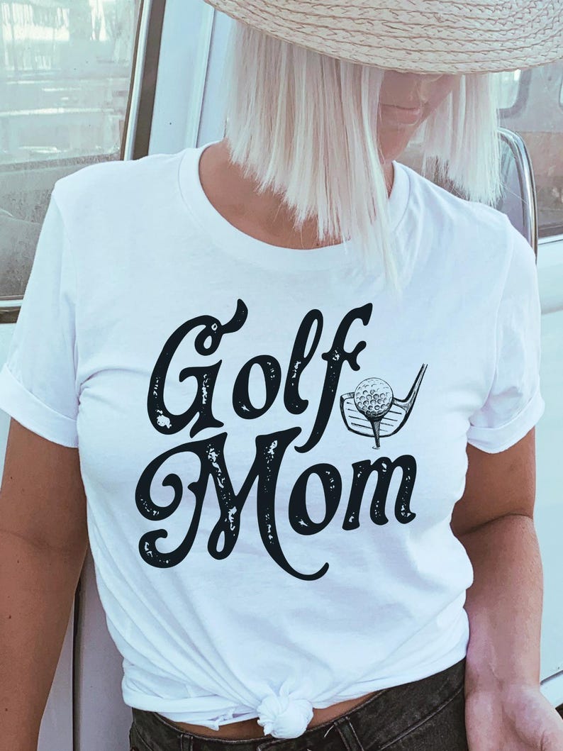 Pu&ograve; includere: Maglietta bianca con una grafica nera invecchiata che dice "Golf Mom" con una mazza da golf e una palla.