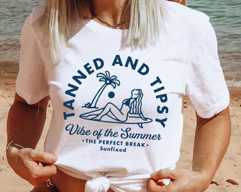 May include: White t-shirt with a blue graphic of a woman on a surfboard with a palm tree and the text "Tanned and Tipsy Vibe of the Summer - The Perfect Break - Sunfixed"