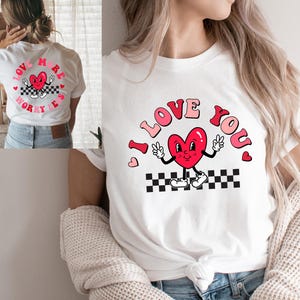 May include: White t-shirt with a retro design featuring a red heart character with the words "I LOVE YOU" in pink and red lettering. The heart is on a black and white checkered pattern.