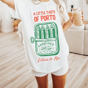 May include: White t-shirt with a graphic of a sardine can, featuring the text "A Little Taste of Porto" in red and green. The sardine can illustration includes the words "Sardines Wild Caught in Olive Oil" and "Delicia do Mar".