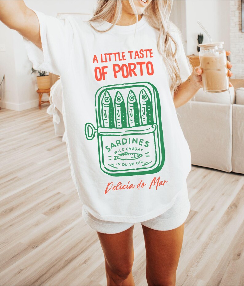 May include: White t-shirt with a graphic of a sardine can, featuring the text "A Little Taste of Porto" in red and green. The sardine can illustration includes the words "Sardines Wild Caught in Olive Oil" and "Delicia do Mar".