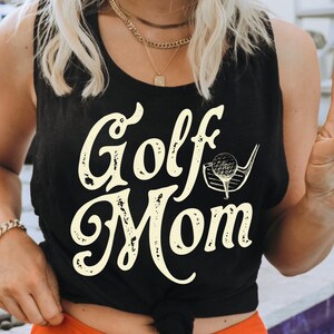 Pu&ograve; includere: Un top nero a spalline con la scritta "Golf Mom" in color crema. Il testo ha un aspetto vintage e usurato. Un bastone da golf e una palla sono raffigurati accanto al testo.