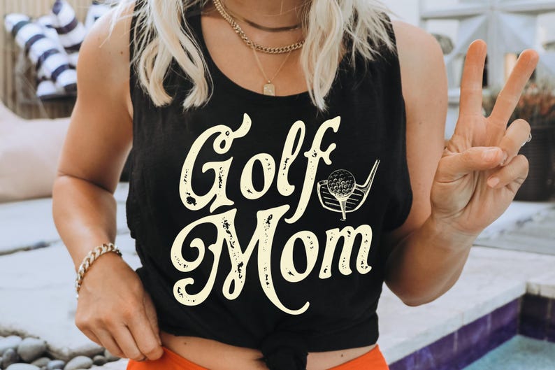 Pu&ograve; includere: Un top nero a spalline con la scritta "Golf Mom" in color crema. Il testo ha un aspetto vintage e usurato. Un bastone da golf e una palla sono raffigurati accanto al testo.