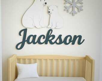 May include: A nursery wall decor arrangement featuring a wooden crib with a white pillow and mattress. Above the crib, there are white polar bear cutouts, a gray snowflake, and the name "Jackson" in teal script. The overall theme is a baby's room.