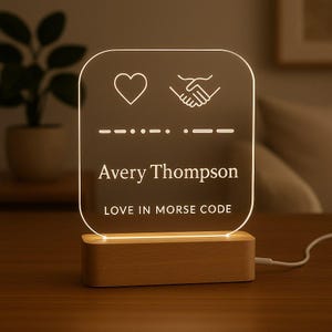 May include: A clear acrylic night light with a wooden base. The light features a heart and handshake icon, Morse code, and the name "Avery Thompson". The text "LOVE IN MORSE CODE" is also displayed.