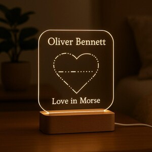 May include: A personalised night light with a wooden base. The clear acrylic panel displays the name "Oliver Bennett" above a heart design in Morse code, with the phrase "Love in Morse" below. The light emits a warm glow.