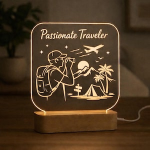 May include: A glowing LED night light with a wooden base. The light features a black background with a white illustration of a traveler with a backpack, mountains, a tent, and a plane. The text "Passionate Traveler" is at the top.