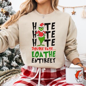 May include: Beige sweatshirt with a Grinch graphic wearing a Santa hat and the text "Hate, Hate, Hate, Double Hate, Loathe Entirely". The person is wearing red and white plaid pajama bottoms and holding a white mug with a Santa Claus design.