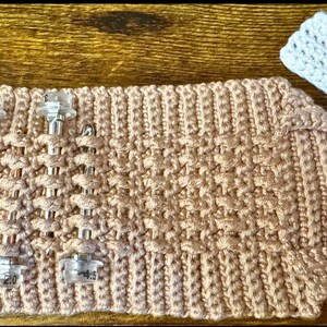 May include: A light brown, hand-knitted cosy with clear plastic clips. The cosy has a textured pattern and a matching white knitted tag attached with a white string. The cosy is designed to hold something.