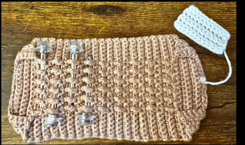 May include: A light brown, hand-knitted cosy with clear plastic clips. The cosy has a textured pattern and a matching white knitted tag attached with a white string. The cosy is designed to hold something.