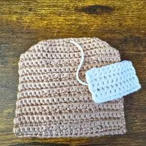 May include: A handmade crocheted tea bag in light brown with a white string and a light blue tag. The tea bag is on a wooden surface. The crochet stitches are clearly visible.