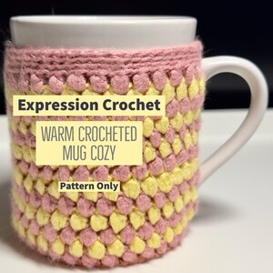 May include: A pink and yellow crocheted mug cosy. The cosy is made with a textured stitch and has a pattern of alternating pink and yellow squares. The text "Expression Crochet" and "Warm Crocheted Mug Cozy" is printed in yellow on the cosy. The text "Pattern Only" is printed in yellow at the bottom of the cosy.