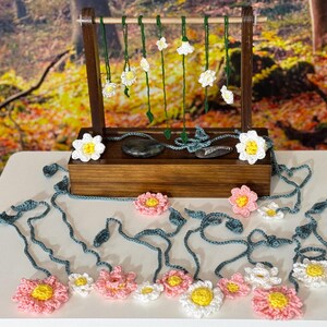 May include: A wooden display stand showcases crocheted flower necklaces with white and pink petals and yellow centres. The necklaces hang from a wooden bar. Additional crocheted flowers and vines are arranged on the surface.
