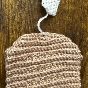 May include: A handmade crochet hat in a light tan colour, with a white crochet leaf attached by a white string. The hat has a rounded shape and a textured surface, suitable for a baby or toddler.