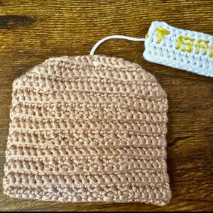 May include: A light brown crocheted tea bag cosy, with a matching crocheted tag that reads "TEA BAG" in yellow thread. The cosy and tag are connected by a white string.